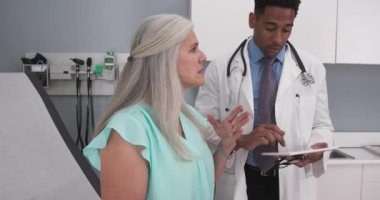 Sevgili yaşlı kadın genç doktora göğüs ağrısını açıklıyor. Siyah milenyum doktoru yüksek teknoloji tablet cihazını kapalı bir sağlık kliniğinde kullanıyor.
