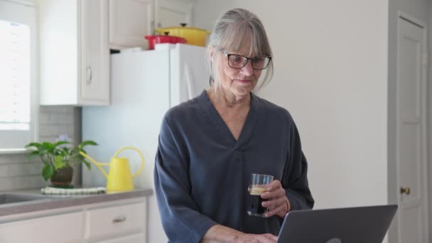 アイスコーヒーを飲みながらキッチンでノートパソコンを使ってシニア白人女性のドリーショット カウンターでパソコンで働くおばあちゃんの中出し スローモーション4K — ストック動画