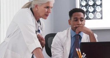 Ofis içinde dizüstü bilgisayar kullanan iki tıp uzmanı birlikte çalışıyor. Genç siyahi doktor hasta dosyalarında yaşlı meslektaşlarına danışmanlık yapıyor.