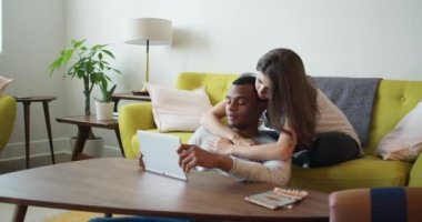 Genç milenyum çifti oturma odalarında tablet bilgisayar kullanıyor. Afrika kökenli Amerikalı ve Kafkasyalı sevgililer mobil cihaz kullanırken konuşuyorlar. 4k Yavaş çekim el bilgisayarı