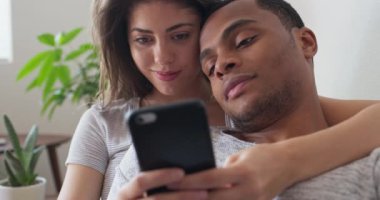 Milenyum çifti bir cep telefonuna bakarken birbirlerini koltukta tutuyorlar. Erkek arkadaş ve kız arkadaş akıllı telefon ekranını paylaşıyor. 4k ağır çekim el bilgisayarı