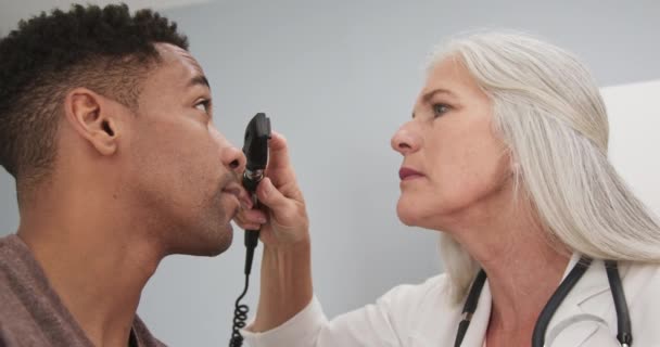 若い黒人男性の患者は 彼の目をシニア女性医師によってチェックした アフリカ系アメリカ人患者の目を調べるために眼科を使用して成熟した女性医師 — ストック動画