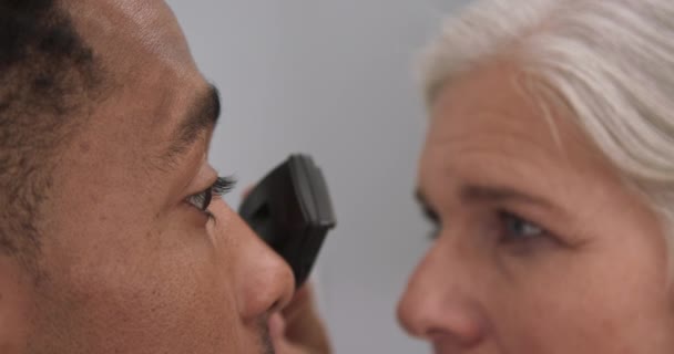 年轻的黑人男性病人由资深女医生检查他的眼睛 用眼科镜检查非洲裔美国人眼睛的成熟女医生 — 图库视频影像