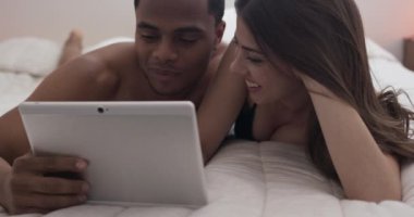 Genç milenyum çifti yatakta yatarken tablet bilgisayarlarında video izliyorlar. Evde cep telefonu kullanan Afro-Amerikalı ve Kafkasyalı kadın ve erkek. 4k ağır çekim el bilgisayarı