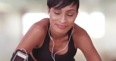 Spor salonunda spor yaparken dinlenen sağlıklı siyah bir kadın. Koşucu kulaklık takıyor ve spor yaparken akıllı telefon kullanıyor. 20 'li yaşlarda gülümseyen Afrikalı Amerikalı..