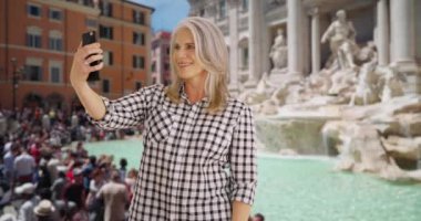 Yaşlı kadın Roma 'daki Trevi çeşmesinde kendi fotoğrafını çekiyor. Kadın İtalya 'da tatildeyken selfie çekmek için akıllı telefon kullanıyor. Kadın telefonda arkadaşlarıyla selfie paylaşır. 4k