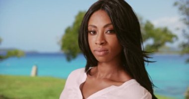 Okyanusun önünde, arkasında yeşil çimenler ve ağaçlar olan güzel siyah bir kadının portresi. 20 'li yaşlarındaki muhteşem Afro-Amerikan kadın tatilde kameraya bakıyor. 4k