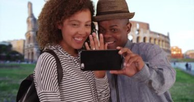 Mutlu nişanlı çift, akıllı telefonlarıyla gülümseyerek ve gülerek fotoğraf çekiyor. Çekici siyah erkek ve kadın, Coliseum 'un önünde poz veriyor. 4k