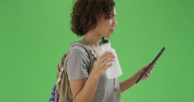 Siyah milenyum kızı yeşil ekranda yürürken tabletini okuyor ve su şişesini içiyor. Anahtar ya da birleşik olmak üzere yeşil ekranda.