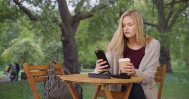 Çekici genç bayan yemyeşil bir parktaki akıllı telefonuyla sosyal medyada geziniyor. Sarışın modern kadın dışarıda kahve içerken telefon kullanır. 4k