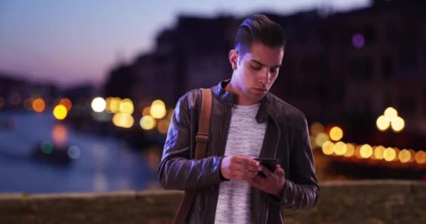 スタイリッシュなヒスパニック千年紀の男は夜にヴェネツィアの橋の上にスマートフォンを見て レザージャケットのラテン男性は ボケライトで夕方に屋外で電話メッセージをチェックします — ストック動画