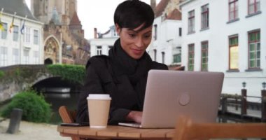Bruges, Belçika 'da güzel bir Afro-Amerikalı kadın serin bir günde dizüstü bilgisayarını kullanıyor. Genç şık siyahi kadın dizüstü bilgisayarında bir e-posta yolluyor. 4k