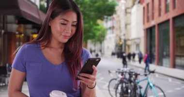 Şehirde cep telefonuyla mesaj atan mutlu Asyalı kadın. Kahve içen ve akıllı telefonda mesajlaşan çekici bir Japon kadın. 4k