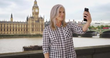 İngiltere 'de seyahat eden olgun bir kadın Big Ben yakınlarında selfie çekiyor. Yaşlı kadın Londra 'da akıllı telefonuyla kendi fotoğrafını çekiyor. Bayan arkadaşlarına telefonda selfie yolluyor. 4k