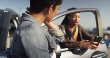 Kız arkadaşlar plajda arabaya yaslanmış cep telefonlarıyla konuşuyor ve mesajlaşıyorlar.