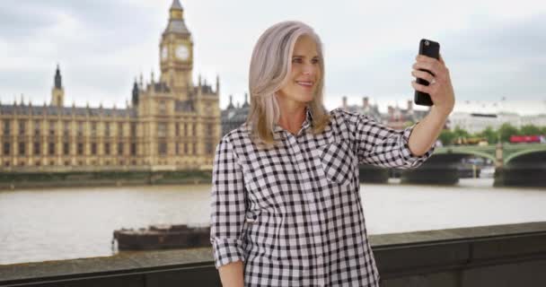 イギリス旅行中の熟女はビッグ ベンの近くで自撮りをする 年上の女性はロンドンのスマートフォンで自分の写真を撮る 女性は友人に電話で自撮りを送る — ストック動画