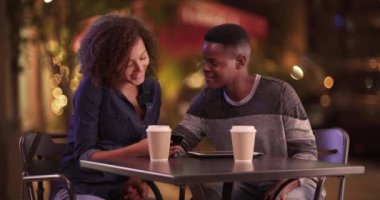 Genç Afro-Amerikan çift geceleri şehirde kahve içerler. Milenyum siyahi erkek ve kadın akıllı telefonları ve tabletleriyle içki içerler. 4k.