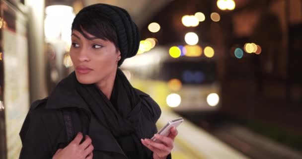 在城市地铁站台上 穿着黑色外套的时尚女人在夜间使用智能手机发短信应用程序 非洲裔美国妇女千年期 在公交车站等候 手持手机 — 图库视频影像