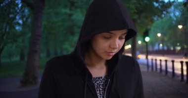 Parkta kapüşonlu kazak giyen Latin bir kadının sıradan portresi. Geceleri sakin ve çekici genç bir kadın kameraya bakıyor. 4k