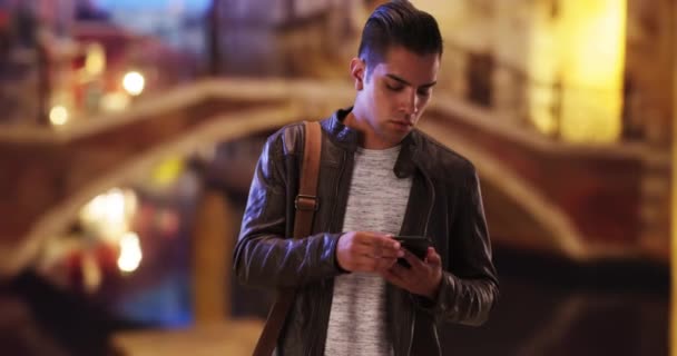 ヒスパニック千年紀の男は夜にヴェネツィアの橋の近くの電話メッセージをチェックします ハンサムなラテン系の男性は夜に外の携帯電話を見て — ストック動画