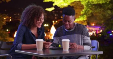 Geceleri dışarıda tablet bilgisayar kullanan çekici siyah erkek ve kadın. Arkadaşlar kahve içmek için buluşuyorlar, modern teknoloji kullanarak ve konuşarak. 4k