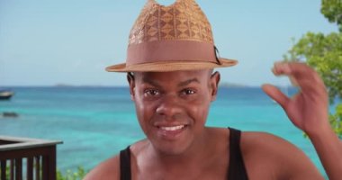 Siyah adam sahildeki bir portre için mutlu bir şekilde poz veriyor. Gülümseyen Afrikalı Amerikalı adam, okyanus kenarında tatilde şapkasıyla oynuyor. 4k.