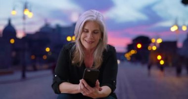 Yaşlı kadın şehirde serin bir akşamda telefonunu kullanıyor. Yaşlı kadın telefonunu kontrol ediyor sonra da şehir sokağındaki kameraya bakıyor. 4k 