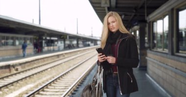 Akıllı telefonunu kullanarak tren istasyonunda sabırla bekleyen güzel bir kadın milenyumu. Trenini bekleyen Kafkasyalı bir kadın kameraya bakıyor. 4k