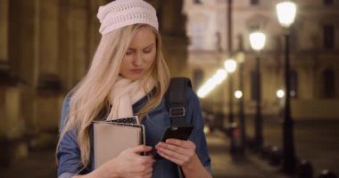 Kafkasyalı üniversite öğrencisi bir gece kampüsün dışında mesaj yazma çalışma grubu. Üniversitede beyaz sarışın kadın akıllı telefon kullanıyor ve sokak lambalarının altında kitap tutuyor. 4k