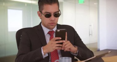 Ofiste cep telefonuyla çalışan genç bir iş adamı. İş adamı akıllı telefon kullanırken içeride güneş gözlüğü takıyor.