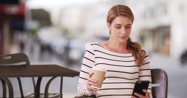 一个随和的女人坐在咖啡店前拿着电话 20多岁的年轻女子在回到工作岗位前享受着休息时间 — 图库视频影像