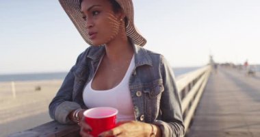 Siyah kadın elinde plastik bardakla iskelede korkuluklara yaslanırken konuşuyor.