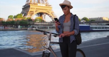 Eyfel Kulesi 'nin yanındaki genç, modern Afrikalı kadın dışarıda telefonunu kullanıyor. Çekici siyahi turist Paris 'te bisiklet sürerken mobil cihazının yönüne bakıyor. 4k
