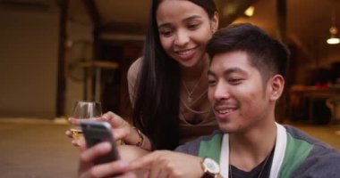 Hoş çoklu etnik arkadaşlar parti girişinde oturup içerken ve gülümserken akıllı telefon kullanıyorlar.