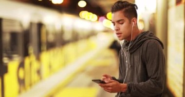 Millennial Latino 'lu adam metro istasyonunda tren beklerken akıllı telefon kullanıyor. İspanyol banliyö, tren istasyonunda müzik dinlemek için cep telefonu kullanıyor. 4k
