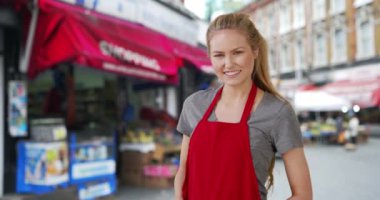 Mutlu beyaz kadın bakkal çalışanı sokaktaki kameraya rahatça poz veriyor. Genç Kafkasyalı işçi çalıştığı mağazanın önünde duruyor. 4k