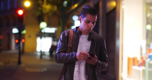ヒスパニック系の千年紀の男は夜に都市部の路上で彼の電話メッセージをチェックします 夕方外のスマートフォンを見ているハンサムなラテン系の男性 — ストック動画