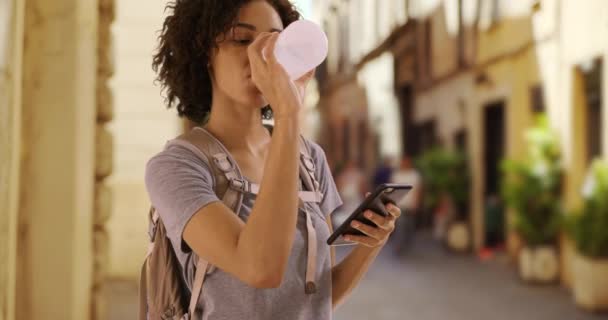 在罗马的小街上 可爱的黑人女性游客一边喝水 一边用手机发短信 年轻的女人在意大利度假时 用手机发短信环顾四周 — 图库视频影像