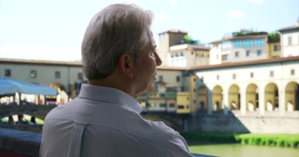イタリアのフィレンツェで見ている間 高齢者の白人男性観光客のポンダー アルノ川沿いの成熟した老人は ヴェッキオ橋 Ponte Vecchio の近くの距離を眺めています — ストック動画