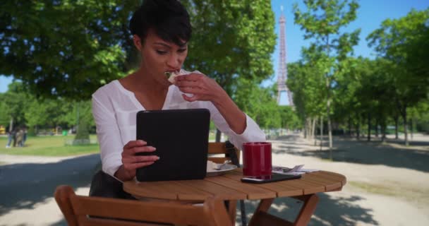彼女のパッド付きの短い髪の民族の美しさは エッフェル塔の近くの屋外テーブルで食事を楽しんでいます パリのアフリカ系アメリカ人女性がタブレットを使用して外に座っている — ストック動画
