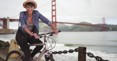 Mutlu gülümseyen siyah kadın San Francisco Golden Gate Köprüsü 'nün önünde bisiklet üstünde oturuyor.