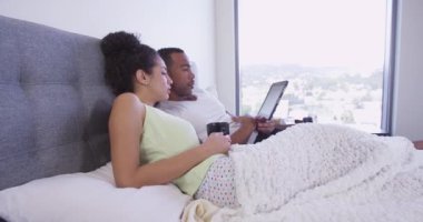 Siyah ve İspanyol çift yatakta oturup tablet bilgisayar kullanıyor ve kahve içiyorlar.