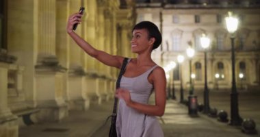 Geceleri ışık altında gülümseyen siyah bir kadın sosyal medyaya koymak için telefonuyla selfie çekiyor. Güzel bir milenyum kadını akıllı telefonuyla kendi fotoğrafını çekiyor. 4k