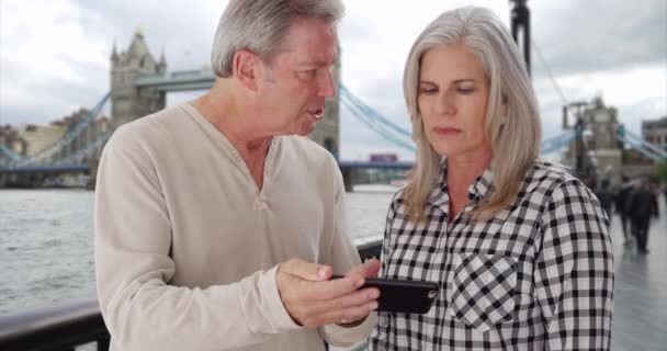 タワーブリッジ近くのスマートフォンで写真を通して成熟した休暇の白いカップルを見てください 古い白人の白人男性と彼の妻はロンドンで休暇中に写真をスワイプします — ストック動画