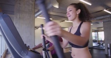 İspanyol kadın gülümsüyor ve eliptik fitness makinesi kullanırken Afrikalı erkek arkadaşla konuşuyor.