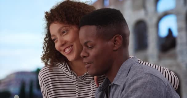 アフリカ系アメリカ人の観光客のカップルは スマートフォンでビデオを見てコロシアムの近くに座っている ローマで黒人男性と女性がおしゃべりし 屋外でリラックスした笑顔 — ストック動画