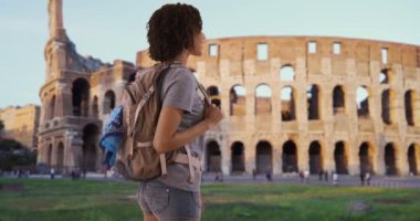 Avrupa 'daki güzel Afrikalı kadın sırt çantaları Roma kolezyumunun keyfini çıkarıyor. Binlerce yıllık mutlu turistin mutlu bir şekilde kameraya gülümseyen portresi. 4k