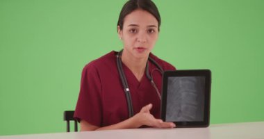 Masada oturmuş tablet bilgisayardaki röntgeni açıklayan İspanyol hemşire ya da doktor. Tıbbi profesyoneller kroma anahtarı veya bileşimi için omurga röntgenini paylaşmak için teknolojiyi kullanıyorlar. 4k