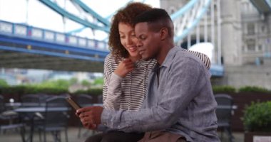 Güzel bir milenyum çifti Tower Bridge yakınlarında akıllı telefon kullanıyor. Mutlu siyah kadın erkek arkadaşının kucağına oturur. 4k