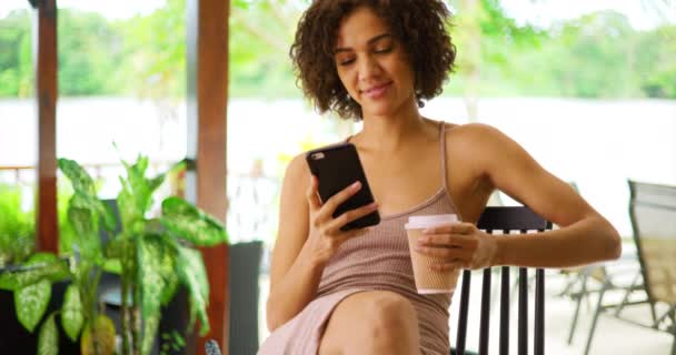 可爱的黑人女人坐在咖啡馆里 喝咖啡和使用智能手机 黑人妇女在网上用手机浏览和在宾馆或度假胜地喝咖啡的肖像 — 图库视频影像